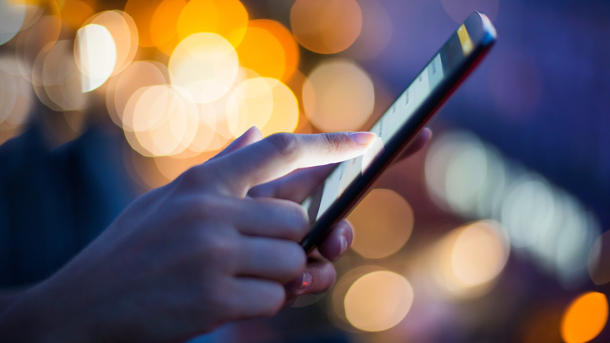 En hand och en mobiltelefon, i kvällsljus med små ljuskällor i oskärpa bakom.