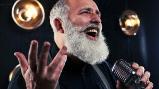 Leende manlig sångare i skägg sjunger i en mikrofon. Lampor i bakgrunden.