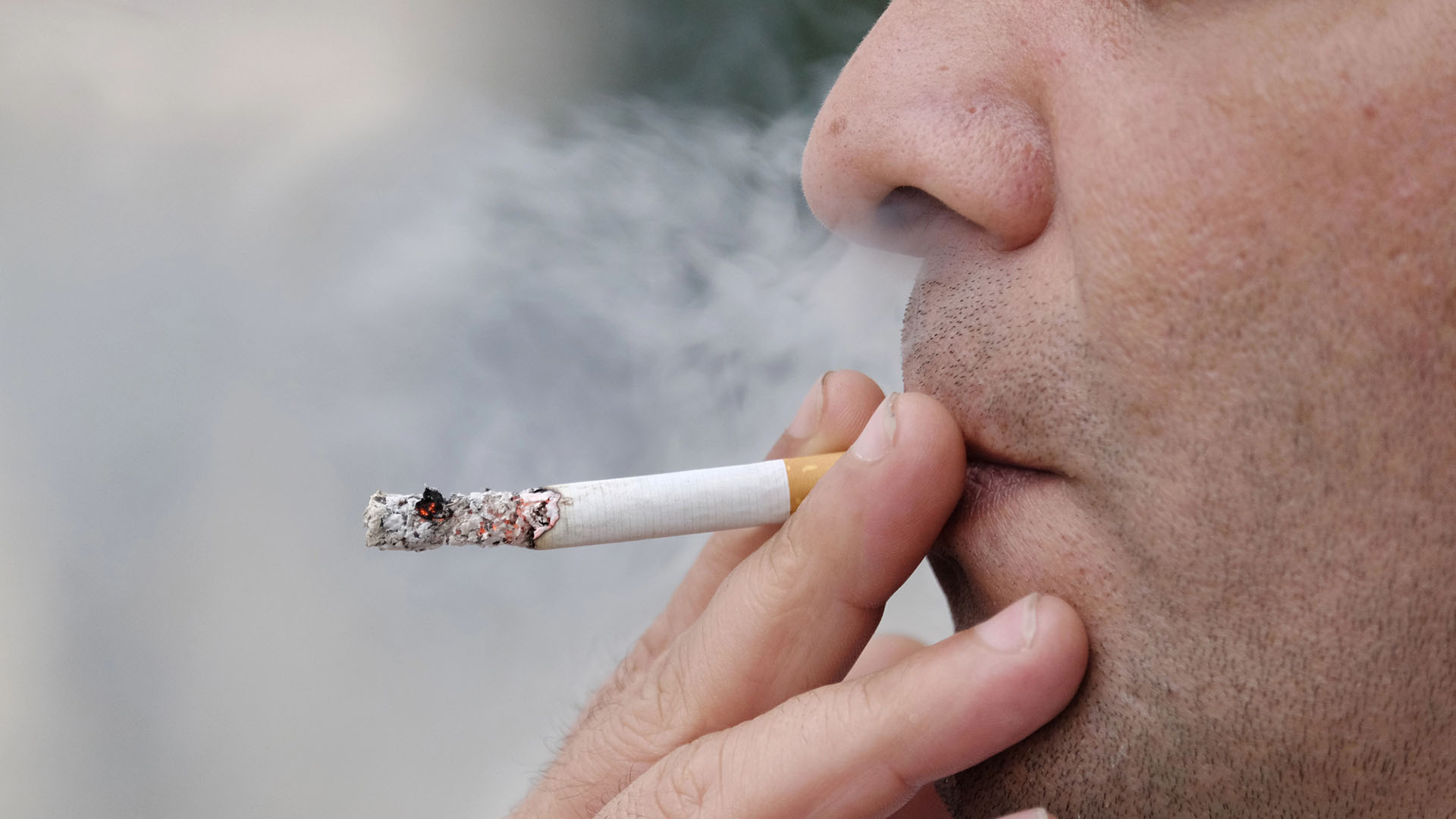 Närbild på en rökare med cigarett i munnen och rök genom näsan.